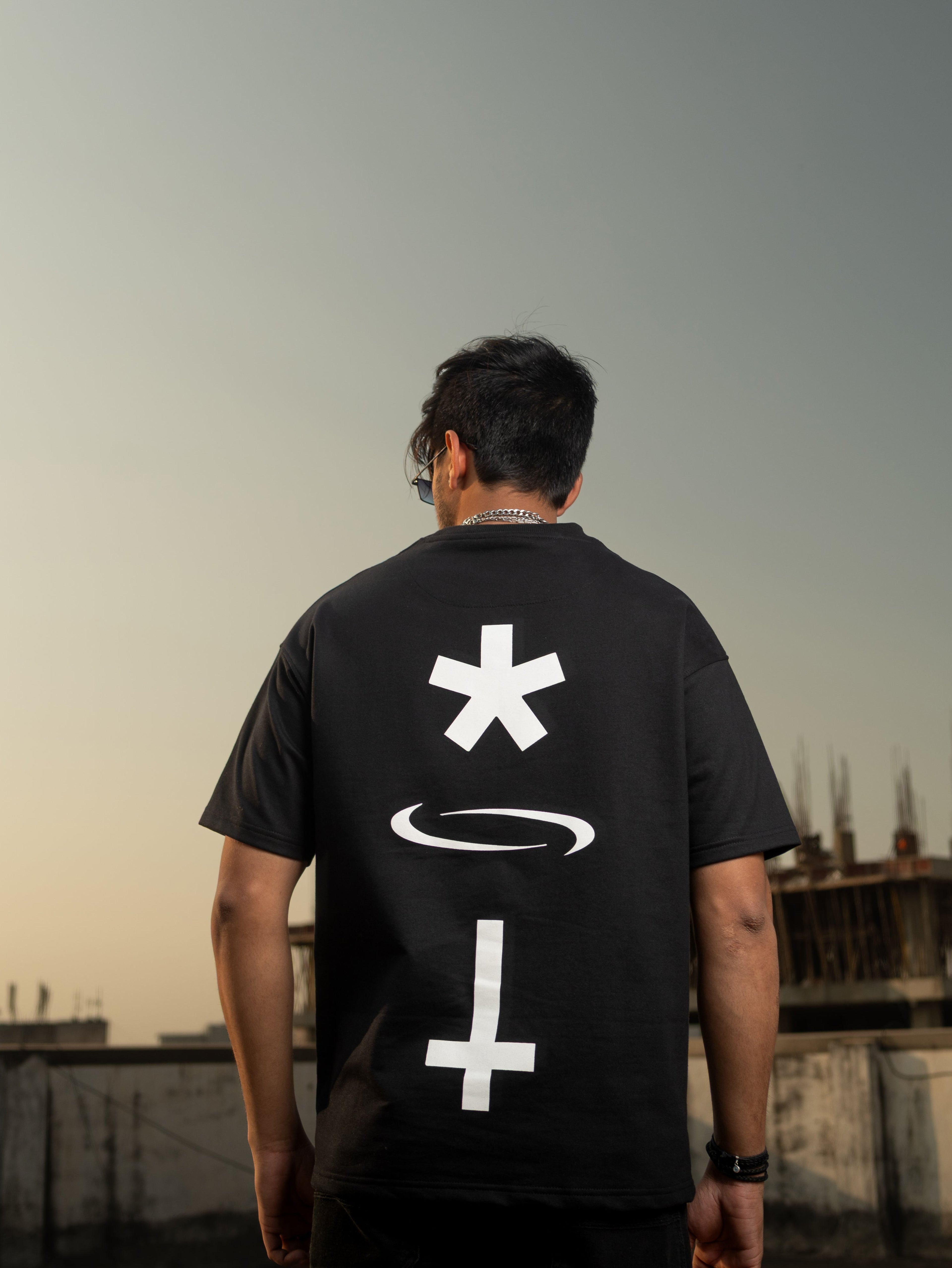 Person wearing a black t-shirt with white symbols on the back, standing on a rooftop with a cityscape in the background.