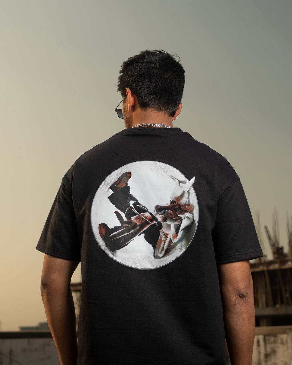 Person wearing a black t-shirt with a graphic design on the back, standing on a rooftop with a cityscape in the background.