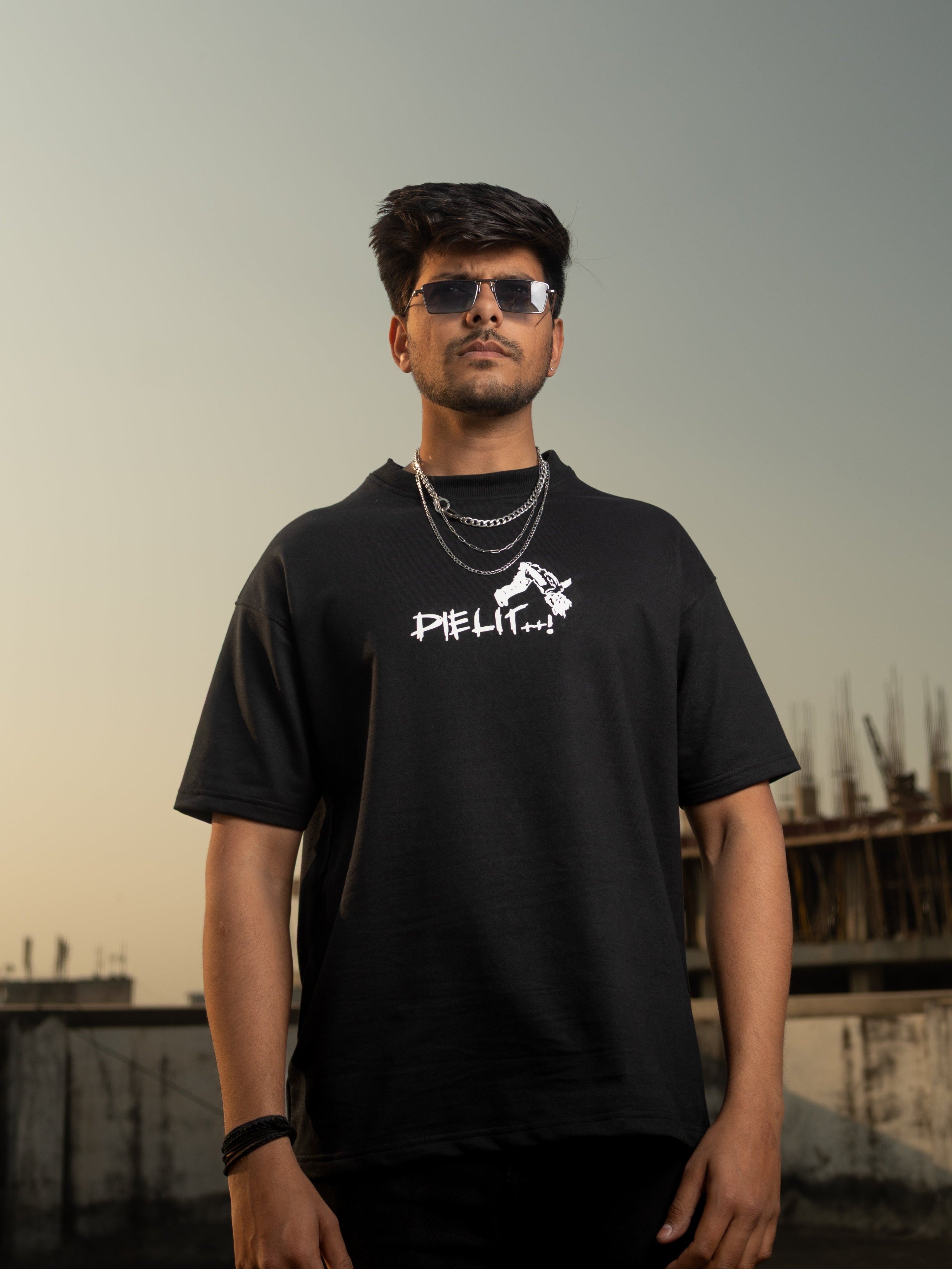 Man wearing a black t-shirt with a logo, standing against an industrial background.